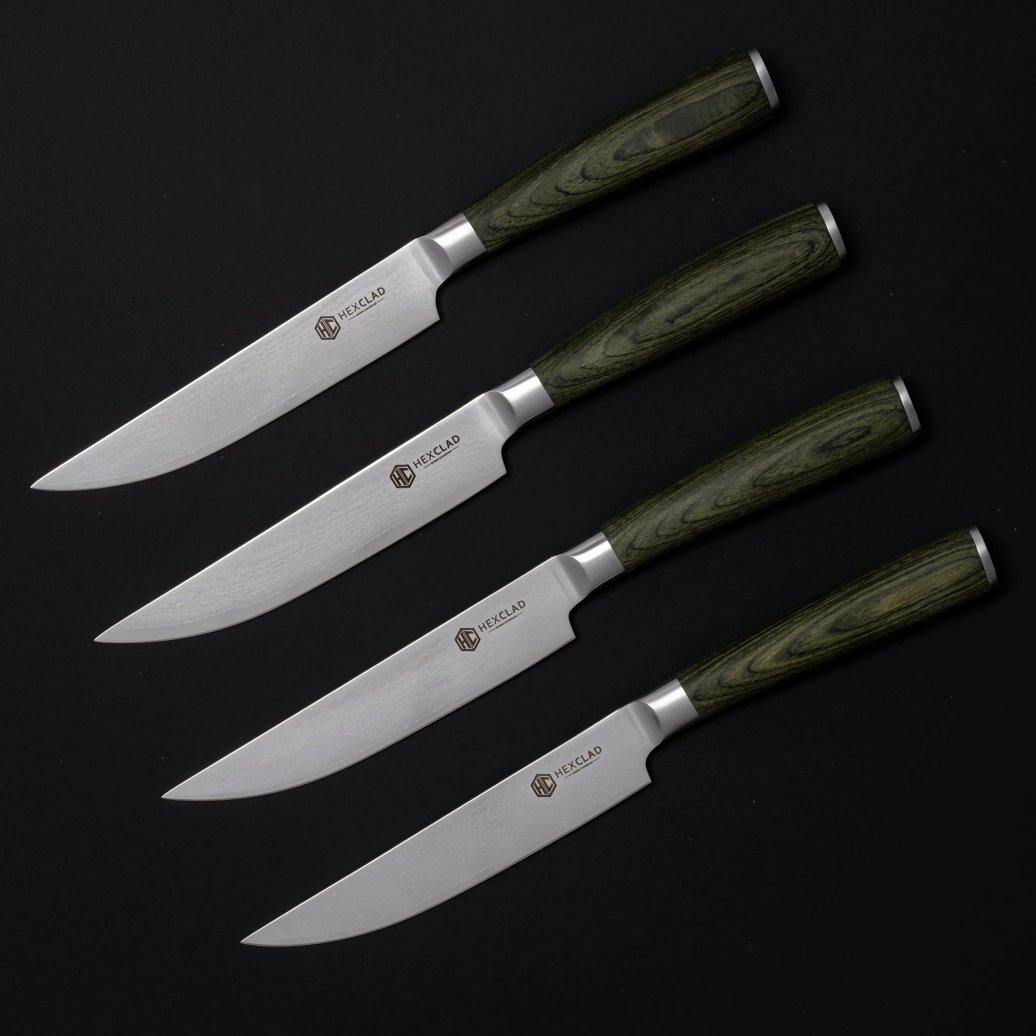 Hexclad biffknivsett. Detalj av fire kniver med skoggrønne pakkatrehåndtak og damaskmønstrede stålblader.