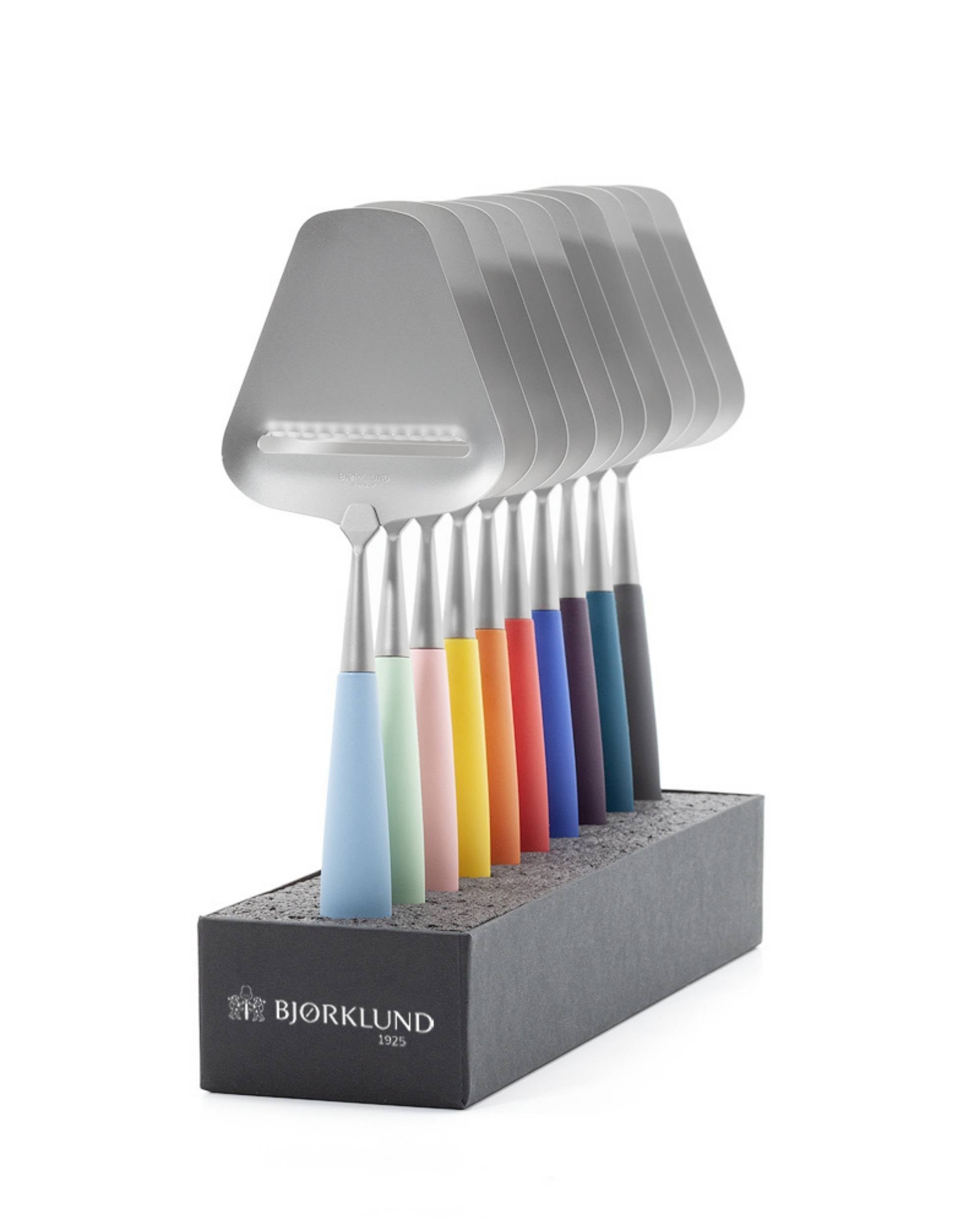 Et sett med 8 Bjørklund Amitto ostehøvler med metallhode, rilleblad, og fargerike plastskaft i en sort holder