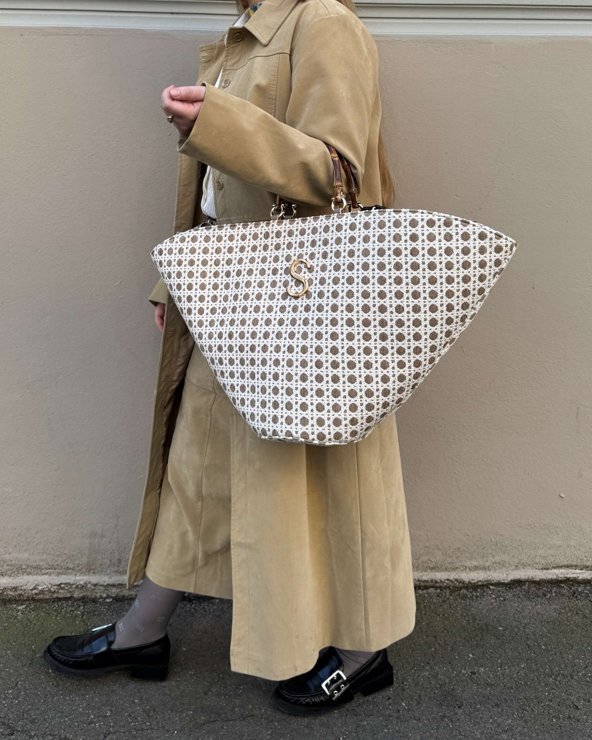 Sorena Capri veske med hvitt og brunt mønster, gullfarget S-logo, bambushåndtak. Holdes av modell med beige jakke.