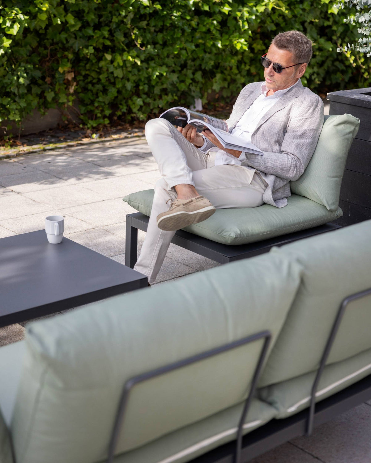 Fiam Domino hagesofa metallramme bakfra, med grønn ryggpute. Mann leser magazine i lignende sofa i bakgrunnen.