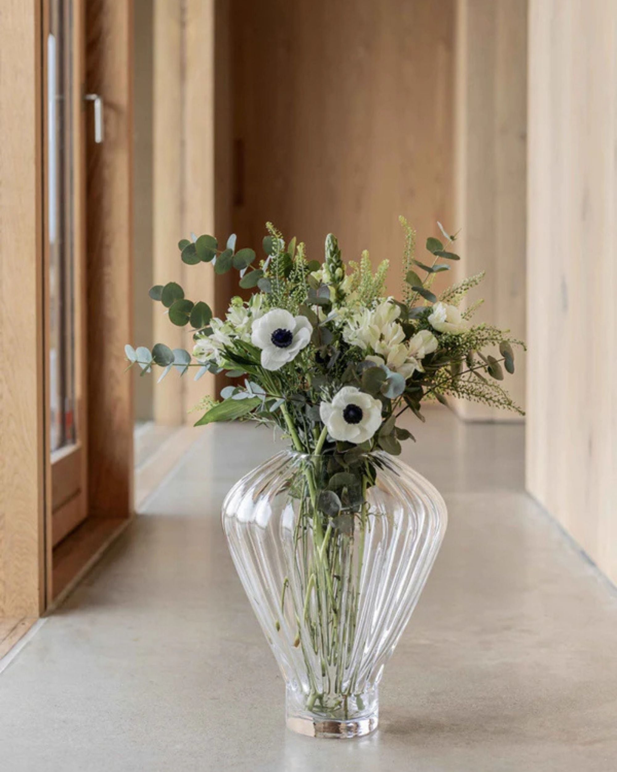 Specktrum Evelyn vase i klart riflet glass med en bukett hvite og grønne blomster, i gang med tregulv