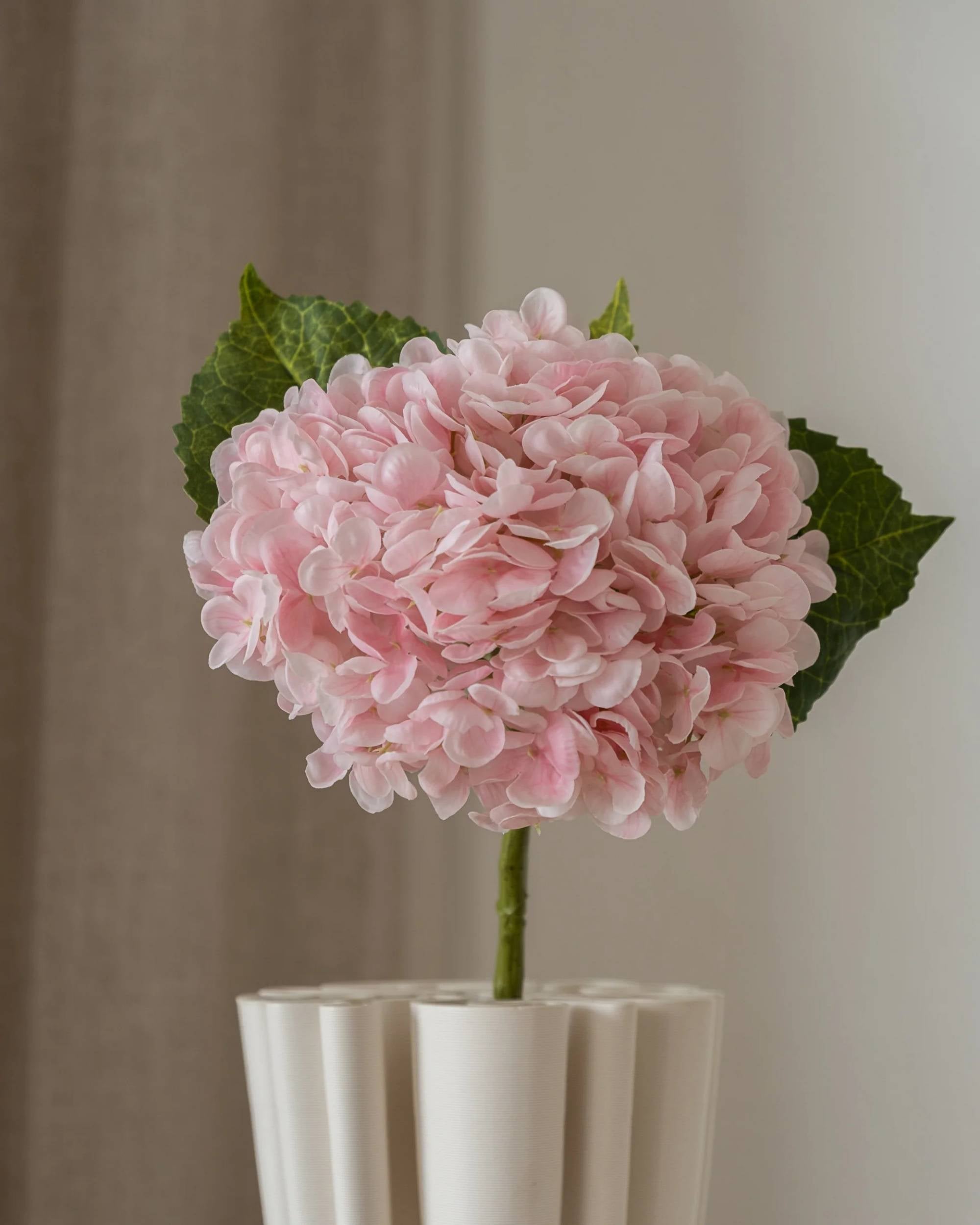 Amarante Hortensia kunstig blomst i lyserosa, detaljerte kronblader og grønne blader, med stengel i hvit vase.