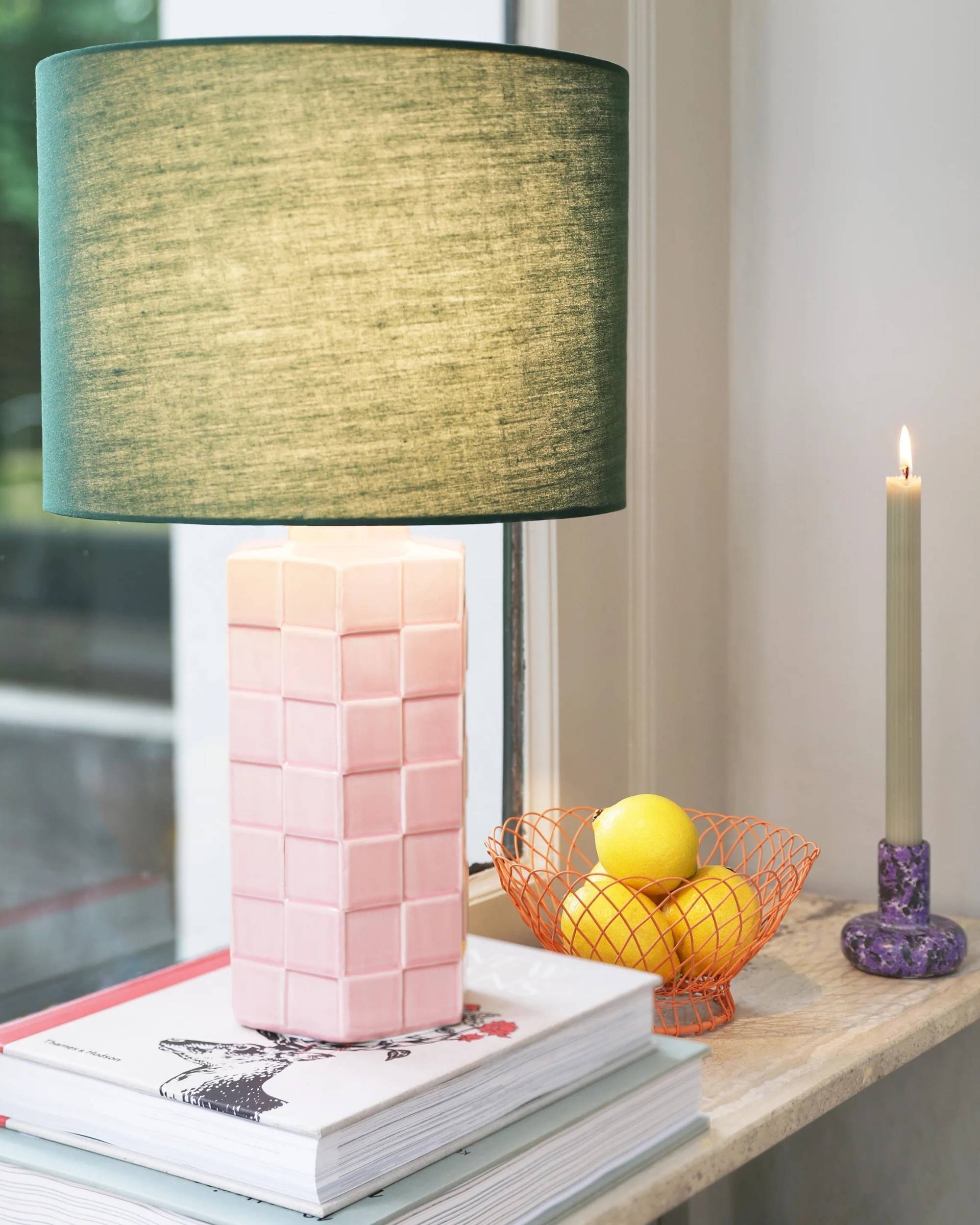 &klevering lampe med rutete, rosa fot og grønn lampeskjerm i stoff, står på bøker ved vindu