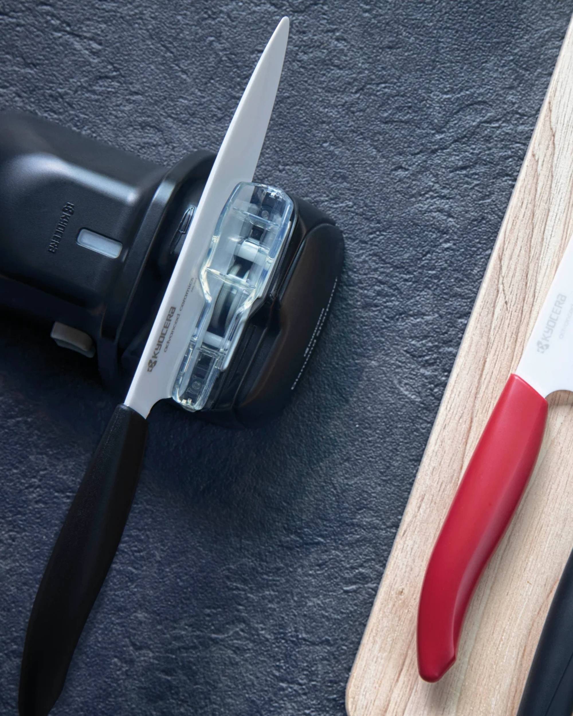 Nærbilde Kyocera batteridrevet knivsliper m/svart kniv, rød knivhåndtak på lyst trebrett.