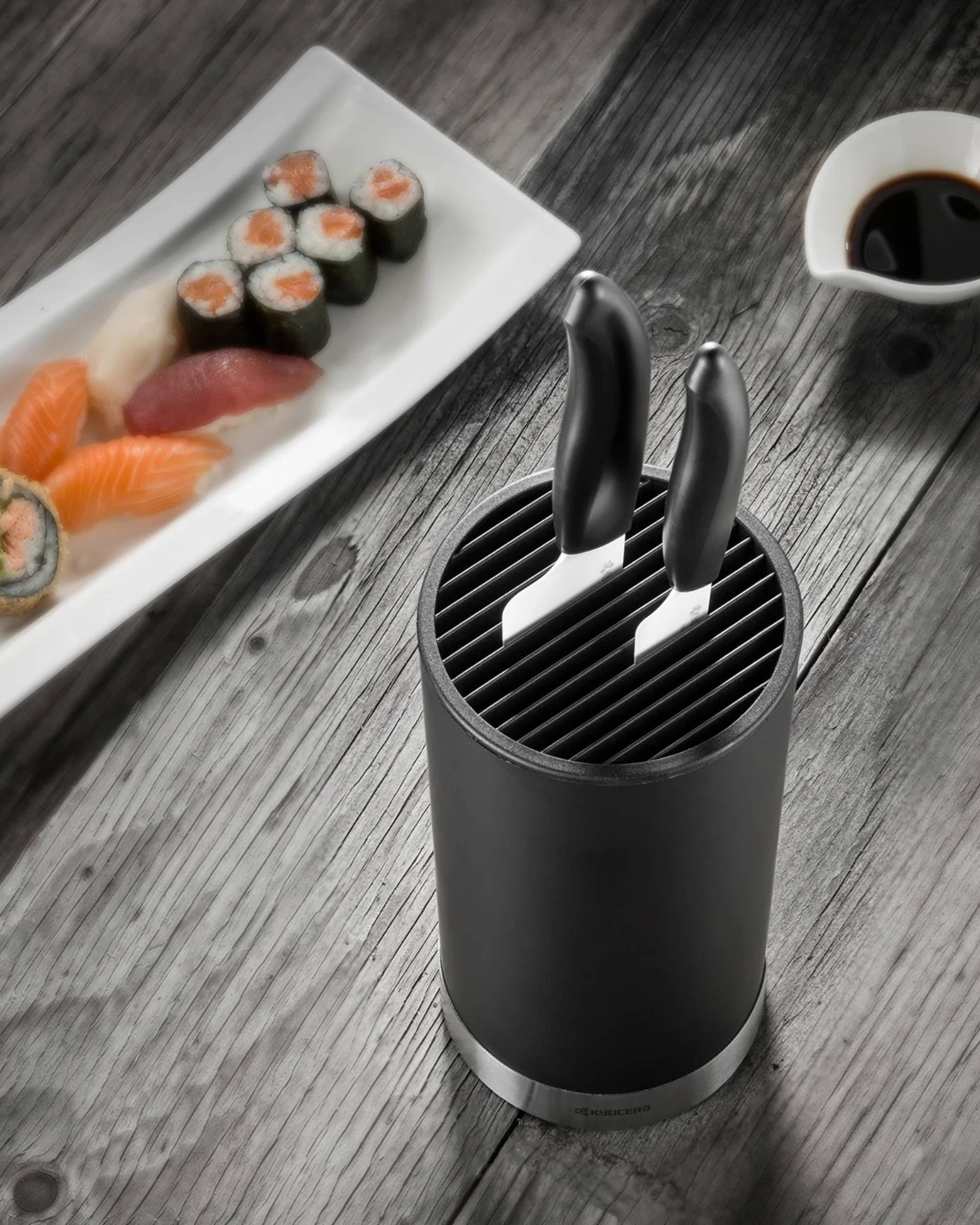 Kyocera knivholder i matt svart med metallbunn, to kokkekniver stikker opp. Sushi og soyasaus i bakgrunn.