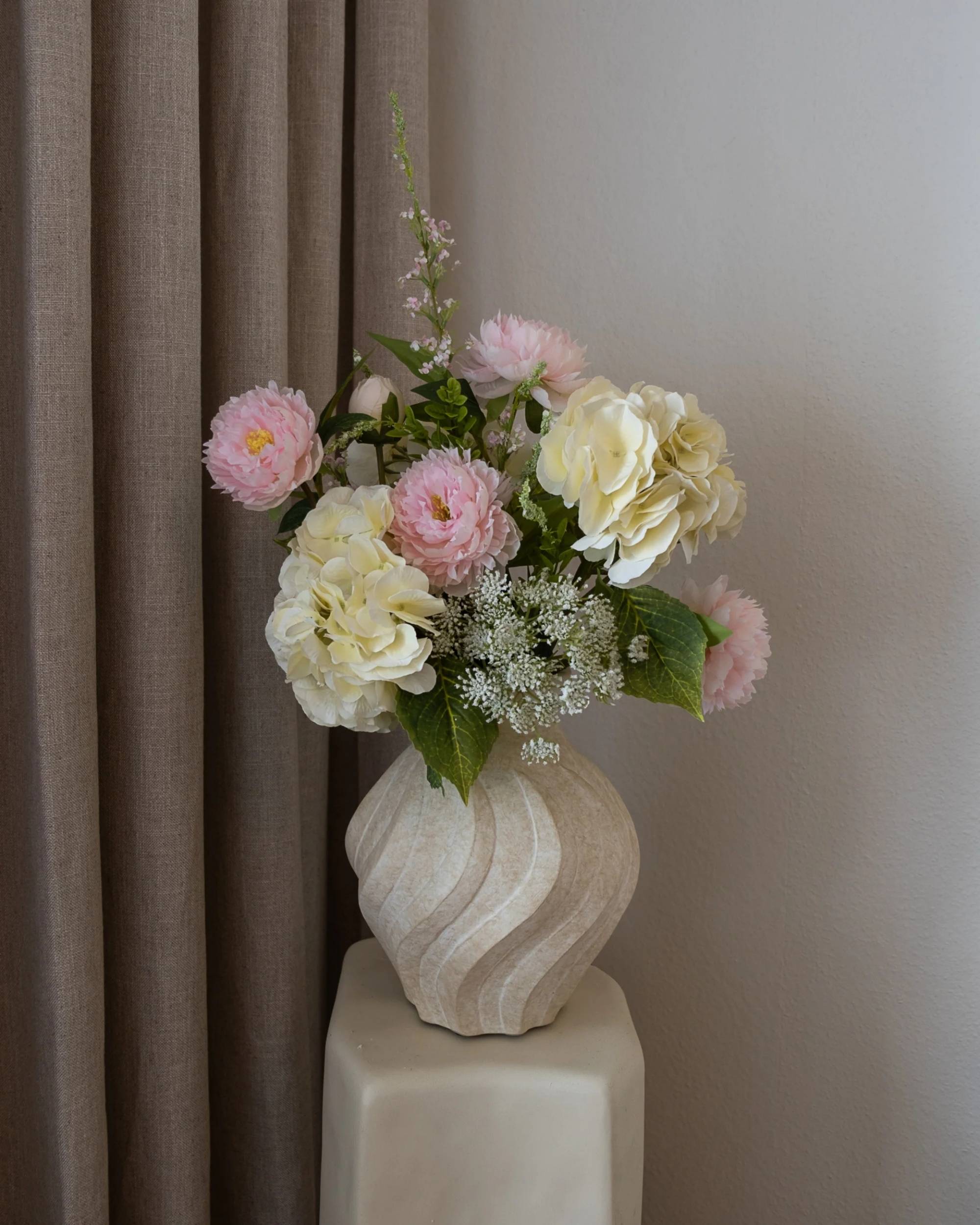 Amarante Strawberry Creme bukett i beige vase, rosa og kremhvite blomster, mot en brun gardin