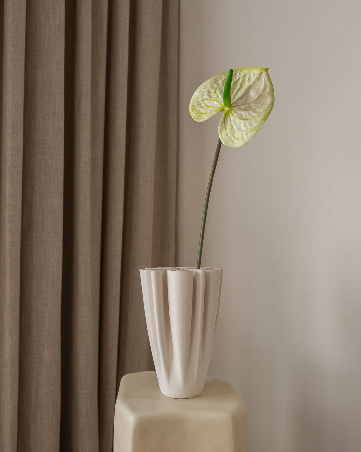 Amarante Anthurium kunstig blomst i hvit vase på pall foran brun gardin og hvit vegg