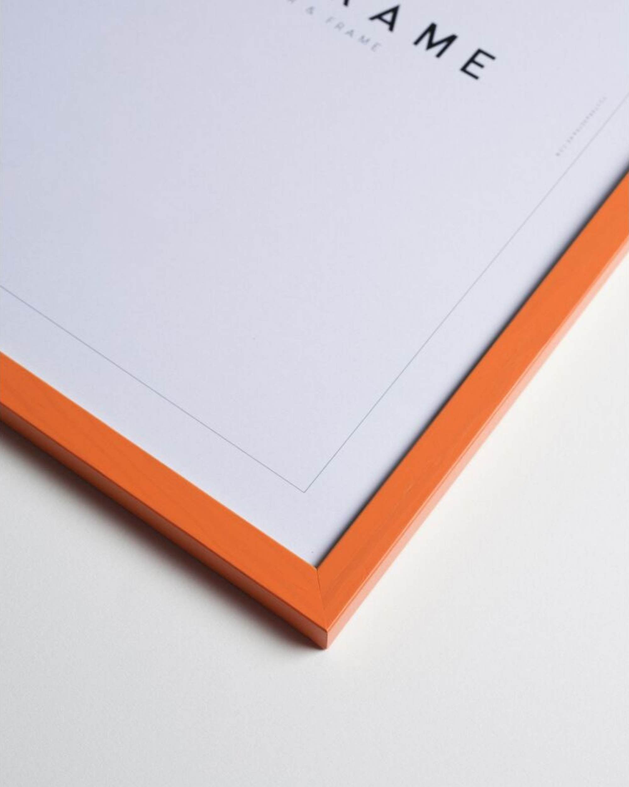 Detalj av oransje bilderamme i heltre fra Poster & Frame, med matt hvit passepartout og del av tekst i bakgrunn.