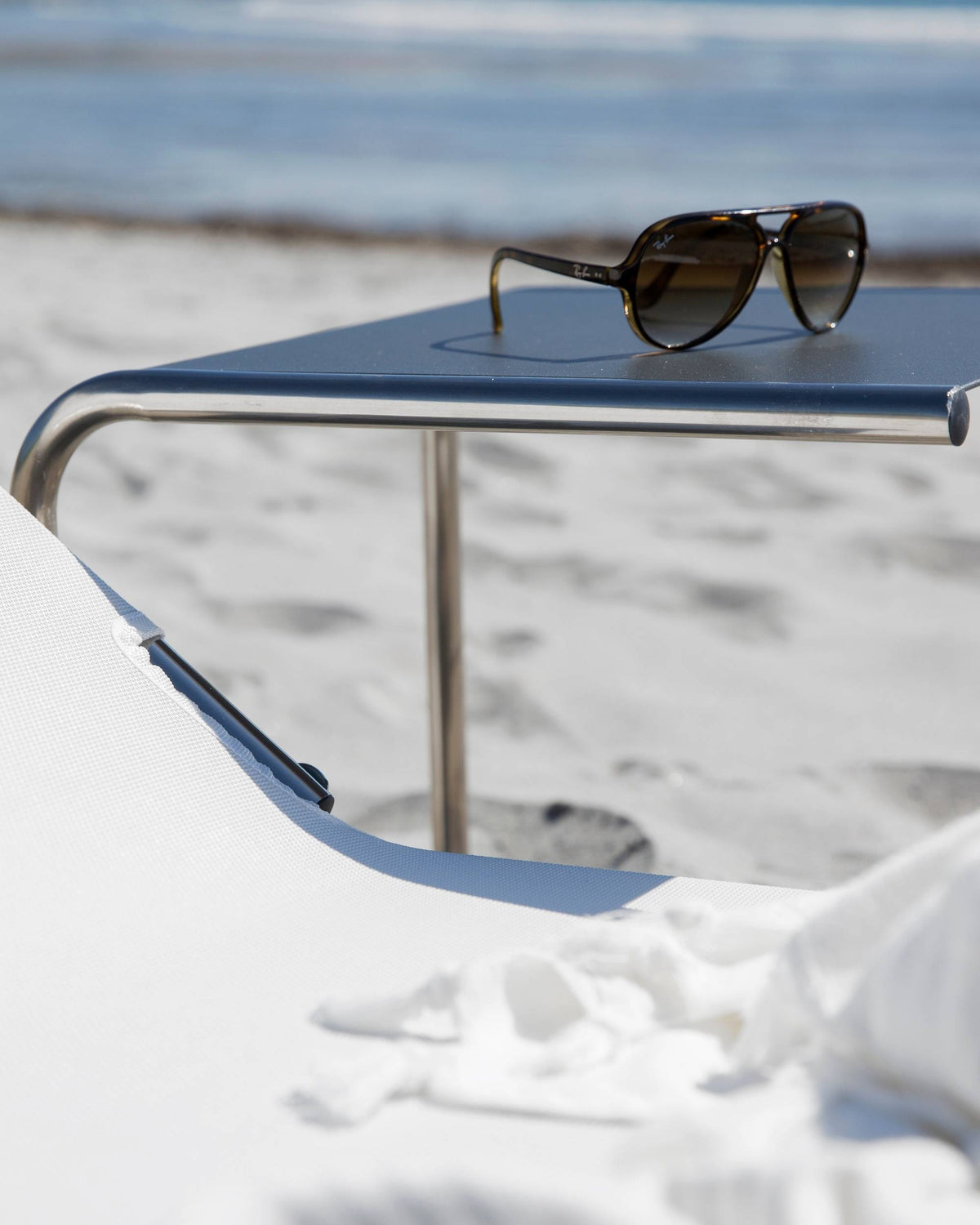 Fiam Club Table hagebord i antrasitt, med solbriller på, og en hvit strandstol i forgrunnen ved havet