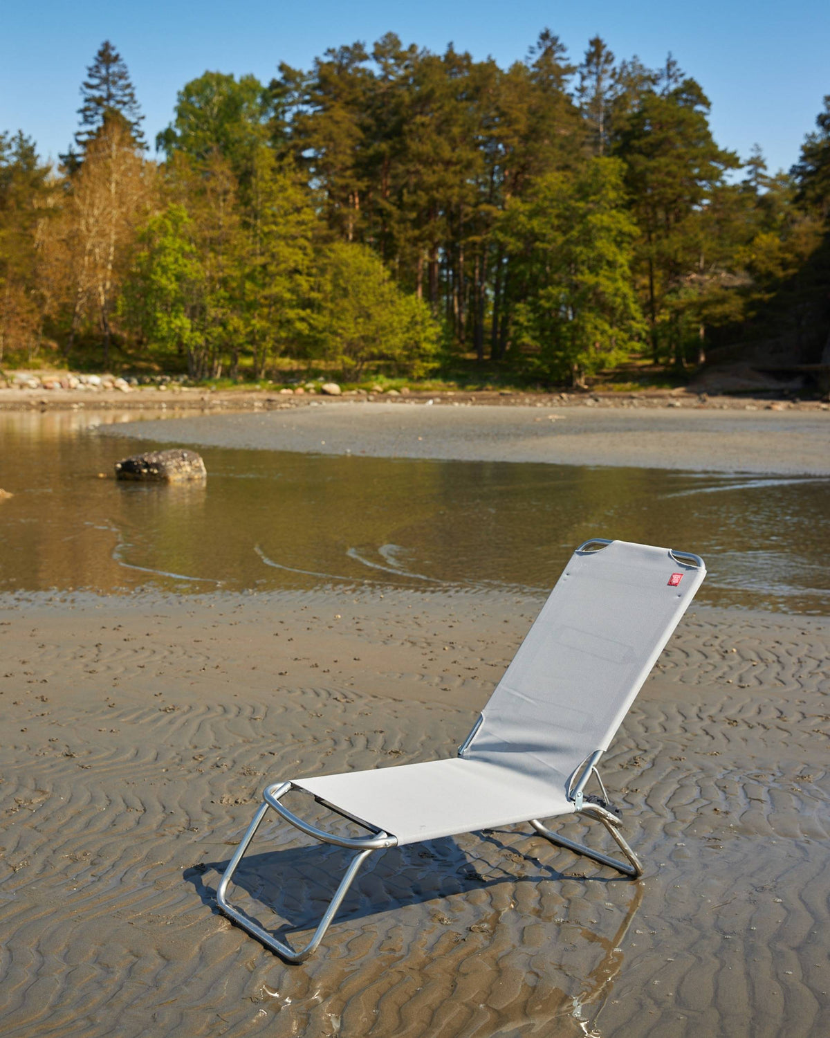 Fiam Jolly strandstol, lys grå texfil og aluminium, justerbar på våt sandstrand med skog i bakgrunnen