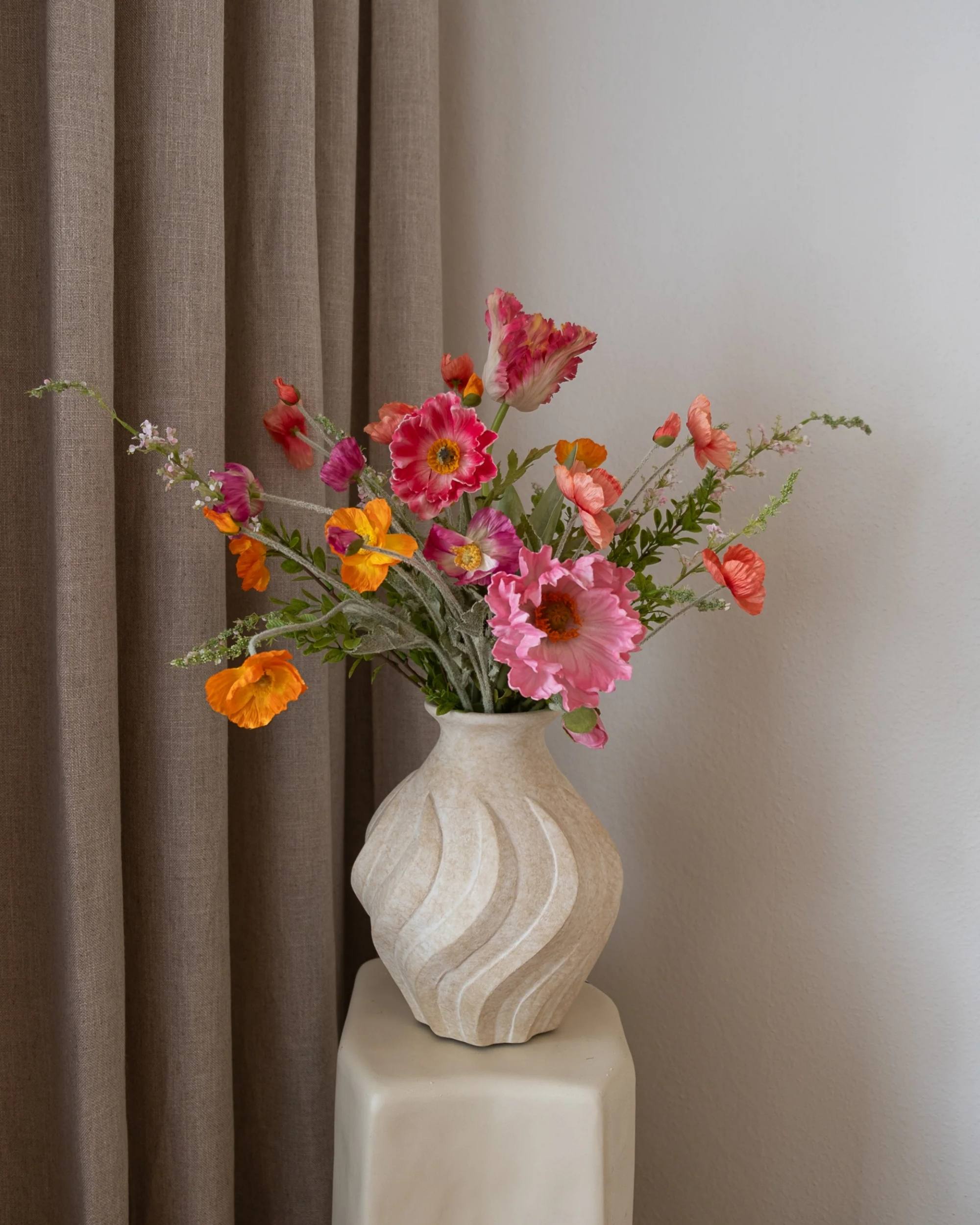 Amarante bukett Pink Lemonade med fargerike naturtro roser i vase på en beige pidestall, mot brun gardin