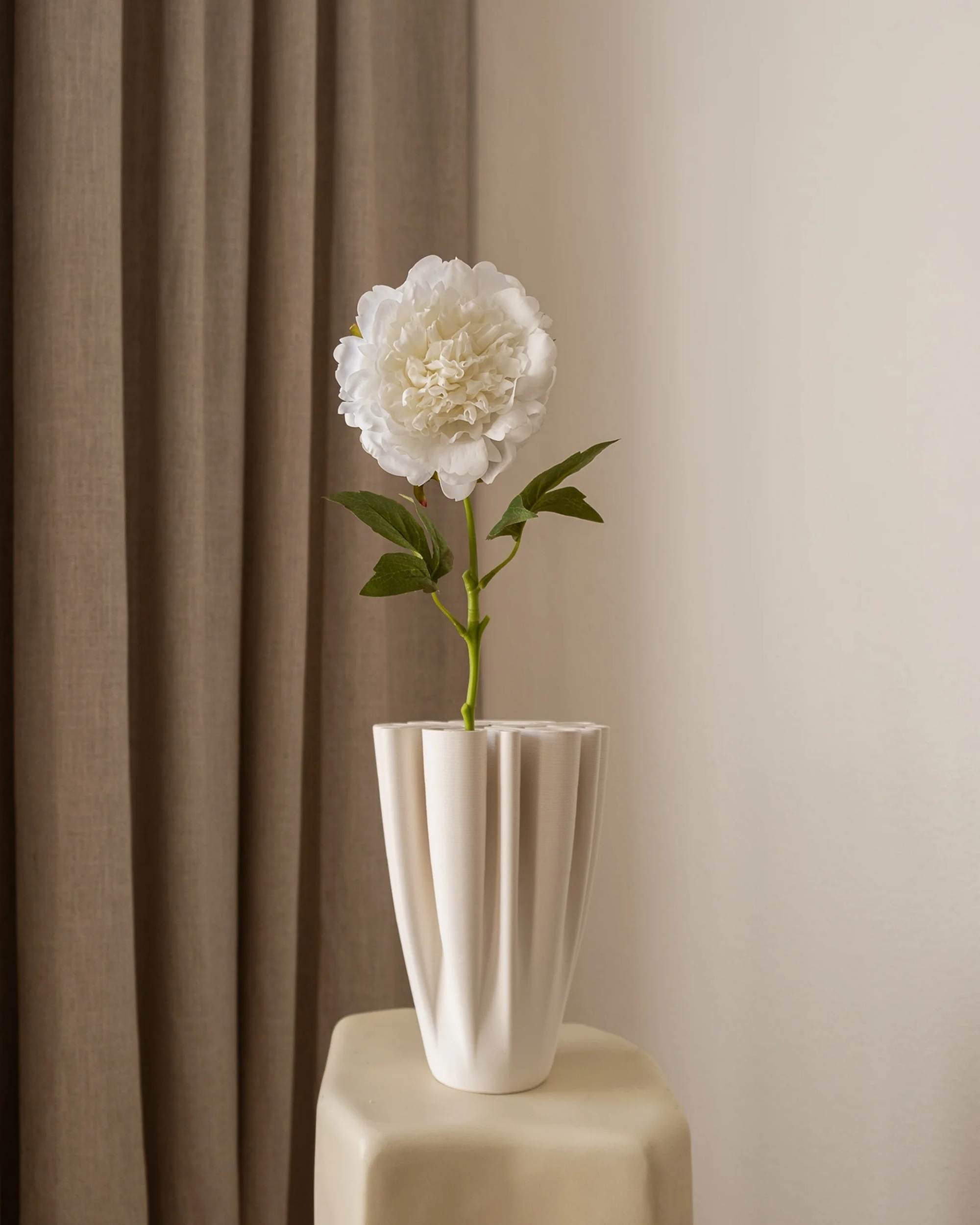 Amarante Peon i Hvit, kunstig blomst med grønne blader, i hvit plissert vase på lyst bord mot gardin