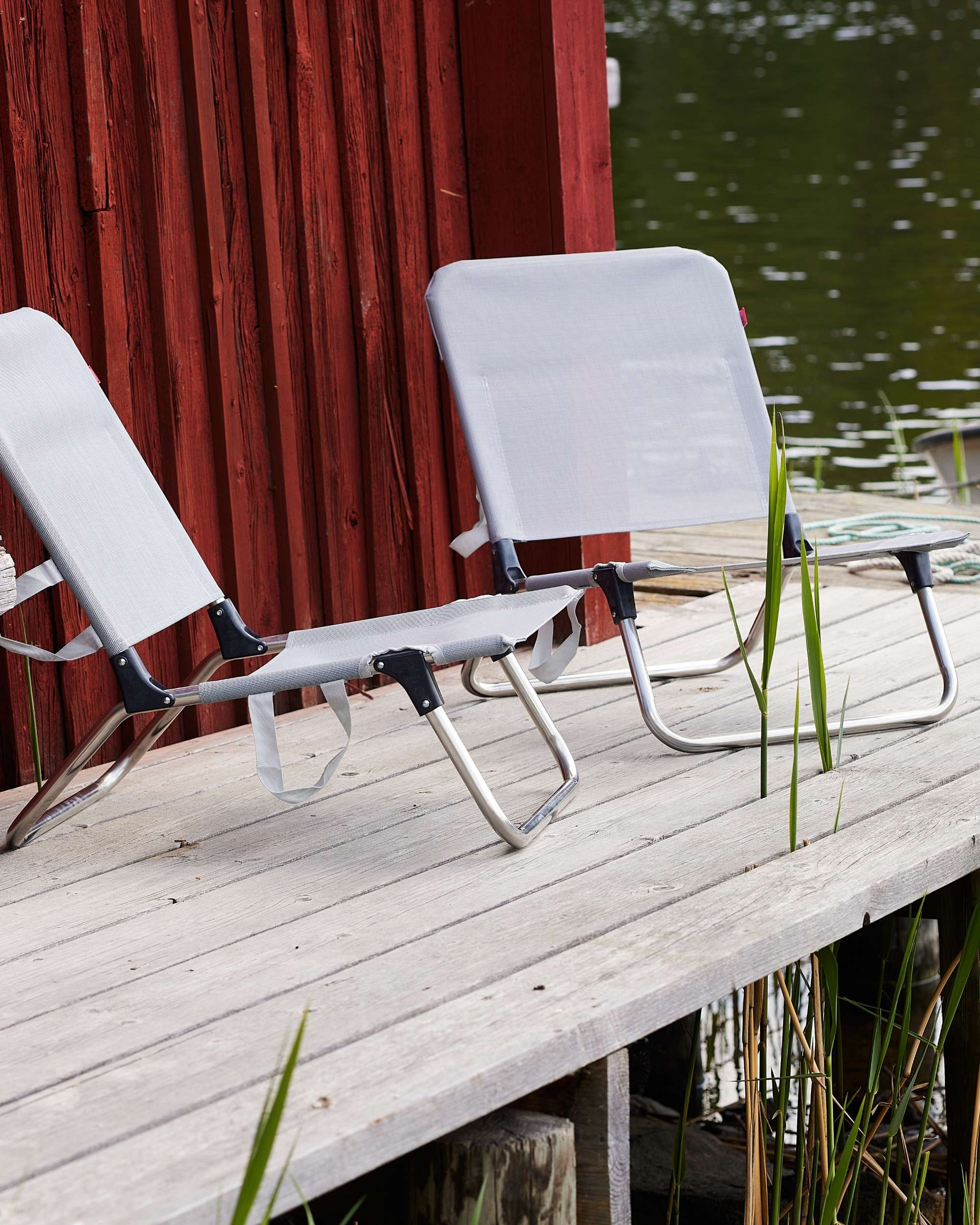 Fiam Quick strandstol detalj: grå tekstilstol med aluminiumsramme på trebrygge ved en rød sjøbod og vann