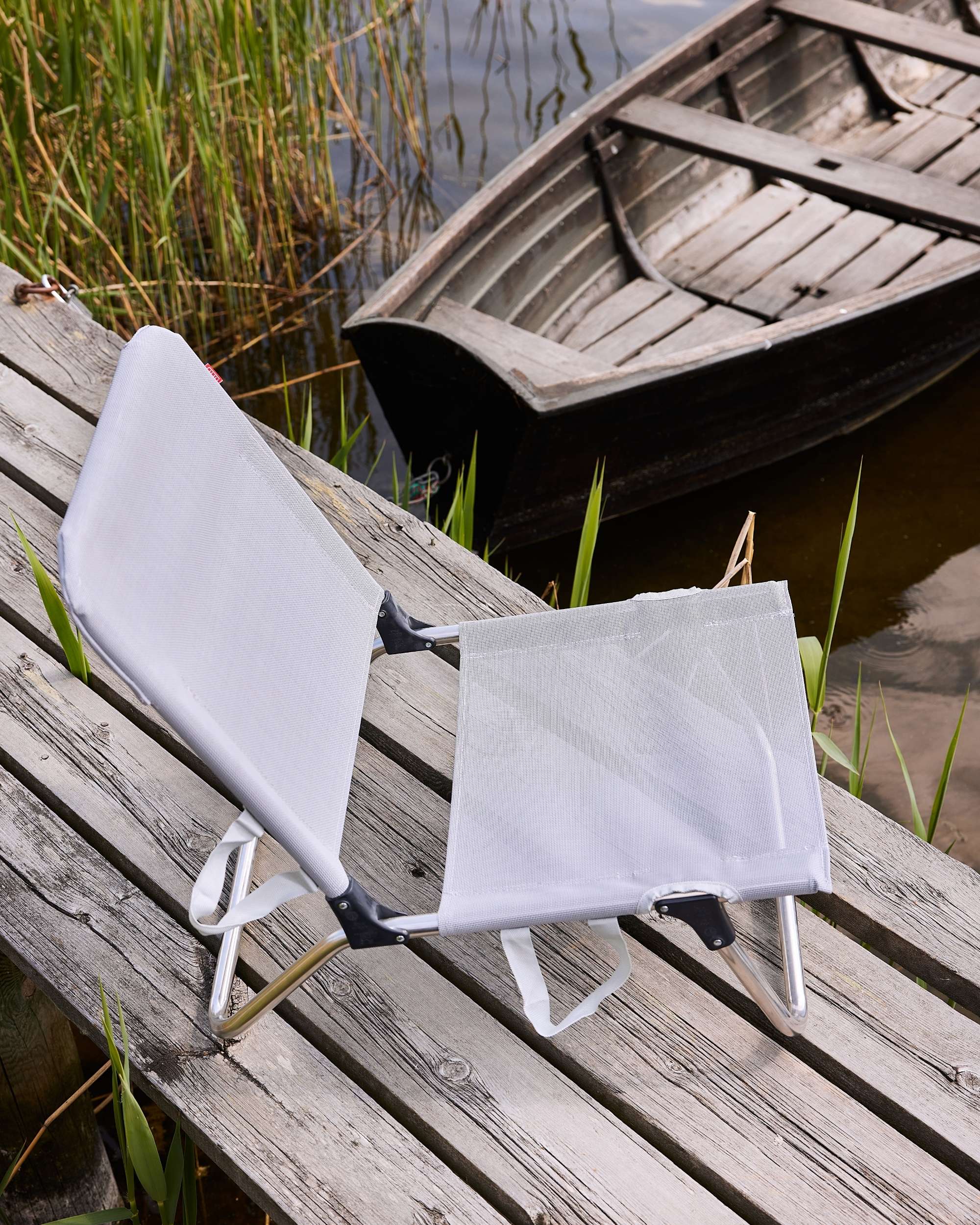 FIAM Quick strandstol i lys grå/hvit tekstil med blank aluminiumsramme, foldet på en trebrygge ved vannet