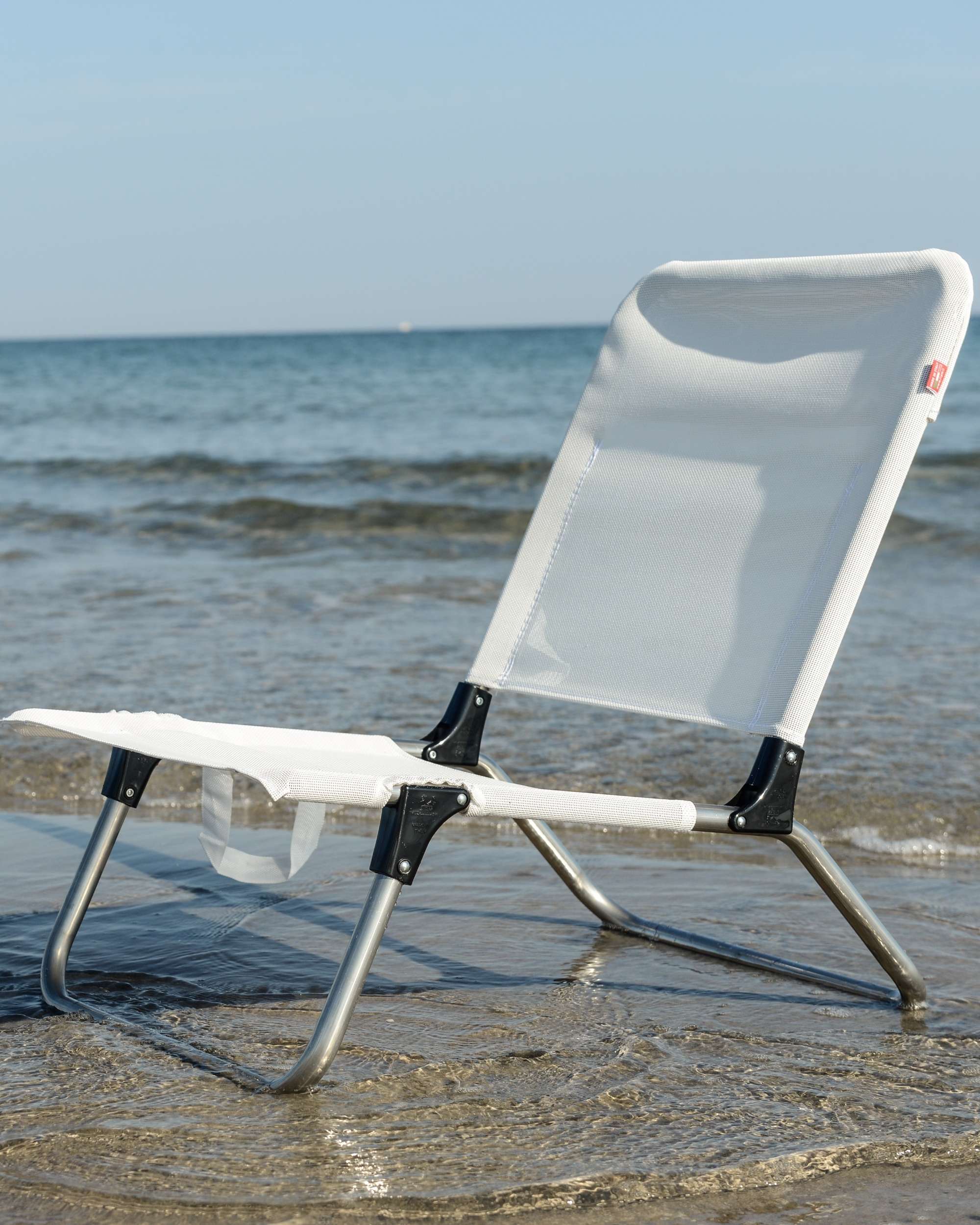 Fiam Quick strandstol i hvit tekstil og aluminium, står delvis i vannkanten på en strand, mot hav og himmel