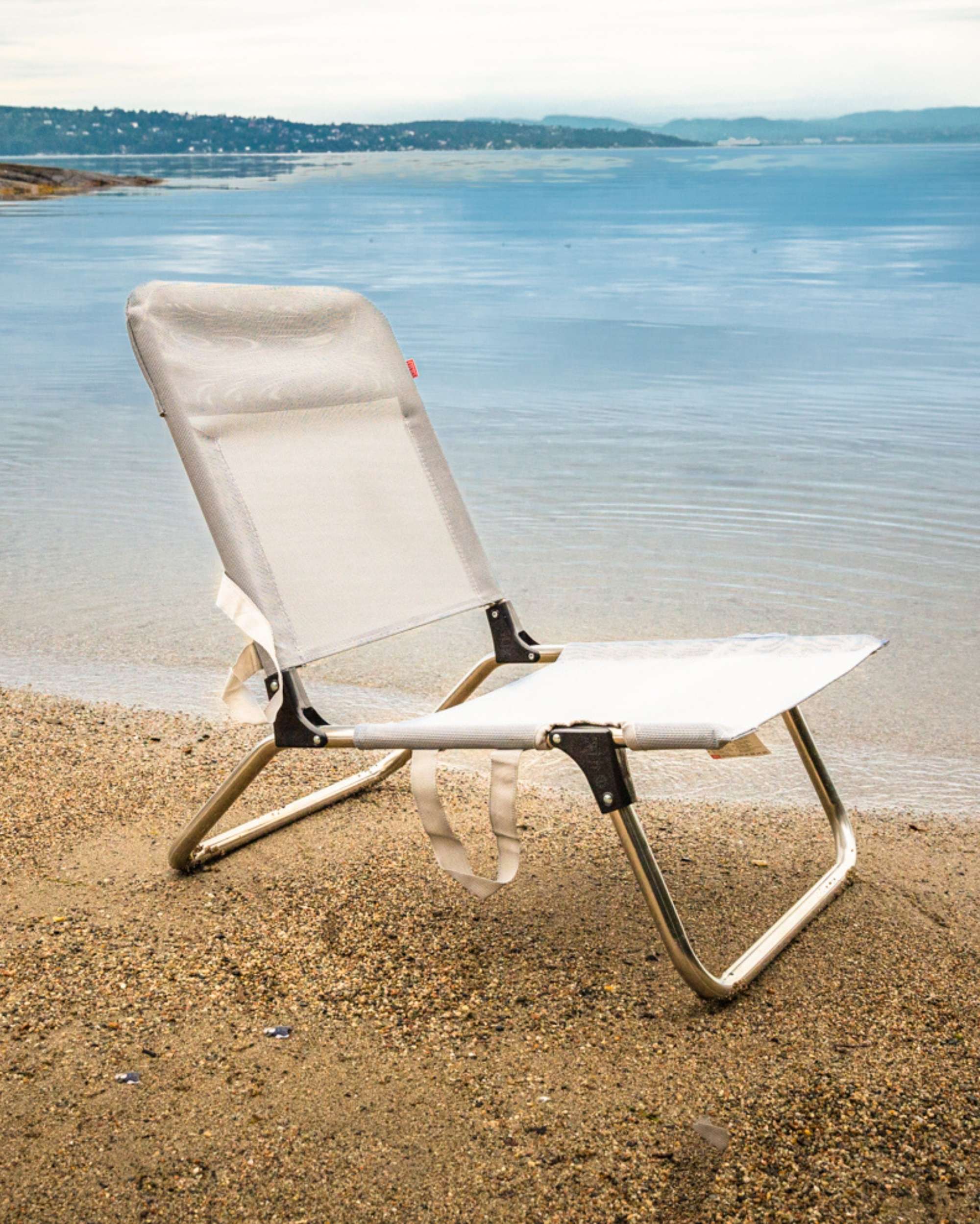 Fiam Quick strandstol med aluminiumsramme og lys grå tekstil, står på sand nær vannet, delvis foldet ut.