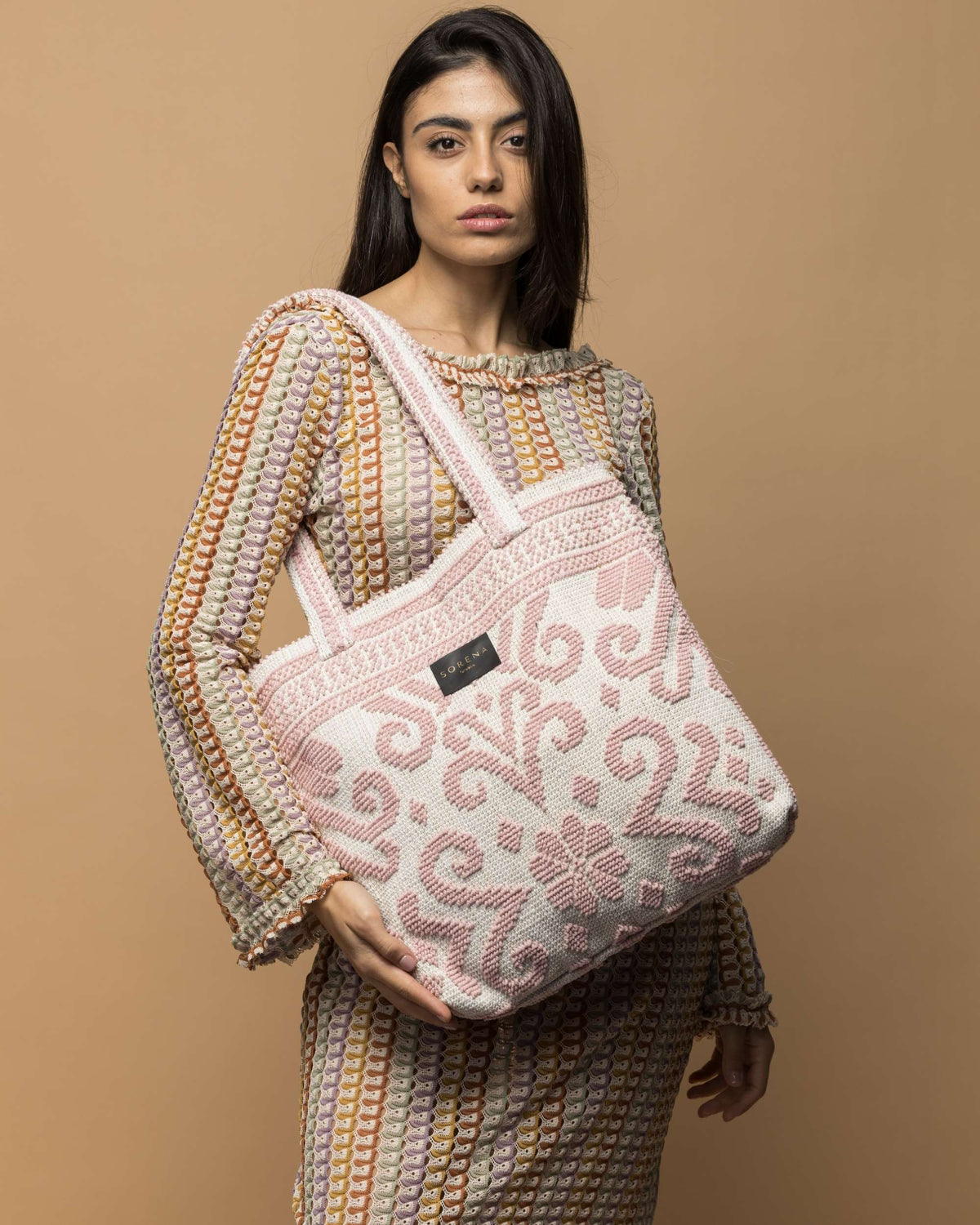 Sorena Caliope BoHo veske i tykt hvitt stoff med rosa heklet mønster, holdt av modell med fluktuøst antrekk